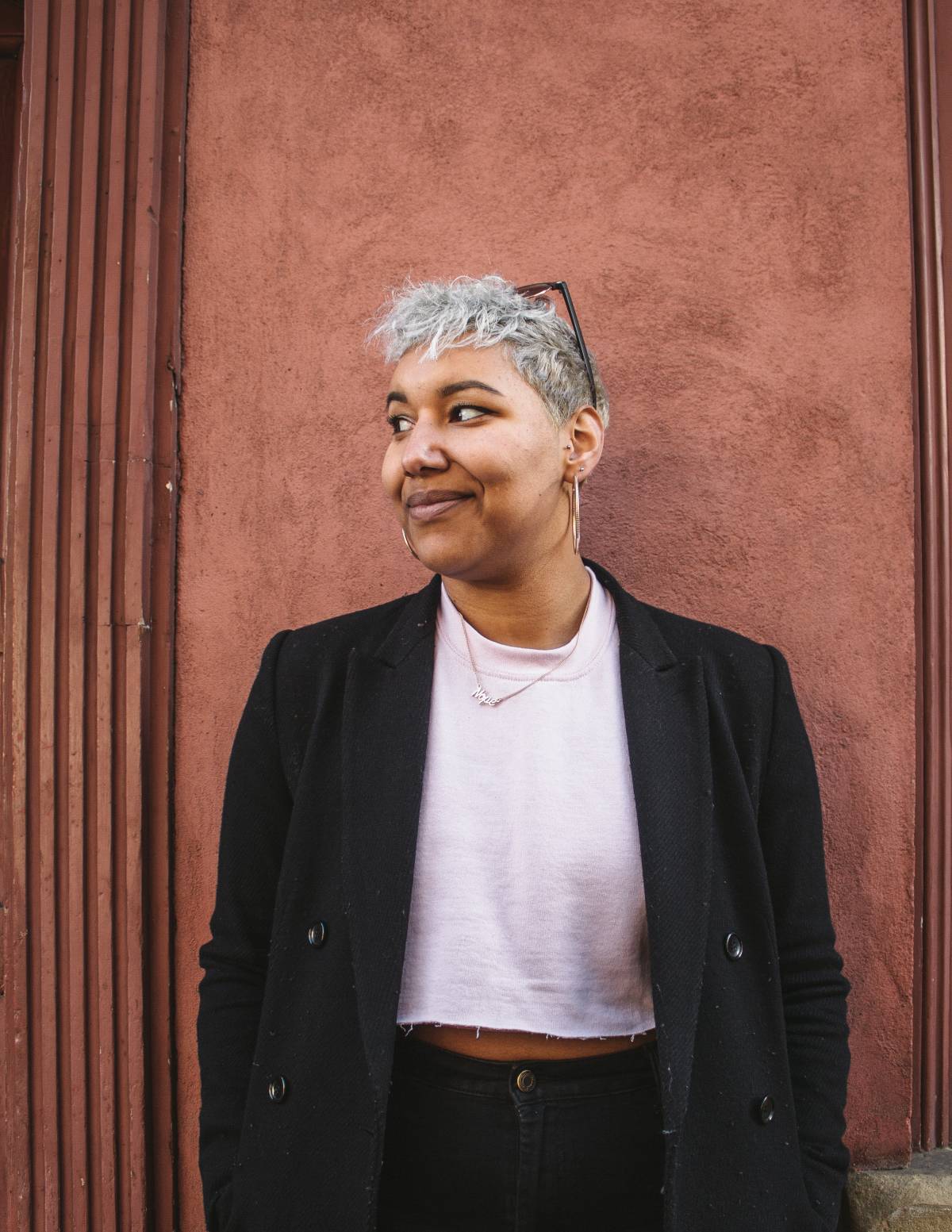 Koko has short silver hair and is wearing a white cropped top underneath a black blazer. She is standing against a dusty pink wall and smiling whilst looking off to the left.