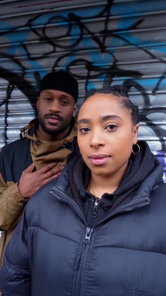 Danny, a black man with a beard and rain mack, and Alizée, a queer mixed-race woman with cainrows and black puffer jacket, stand side by side in front of grey shutters covered in grafitti.