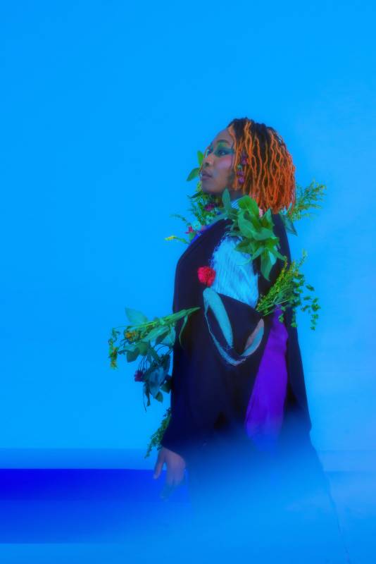 Image shows Ray, a black non binary person with orange, shoulder length locks. They face off to the left, strong and ethereal, on a sky blue background. They are wearing a suit with flowers sprouting from it, ripped at the shoulder revealing a striped shirt underneath.