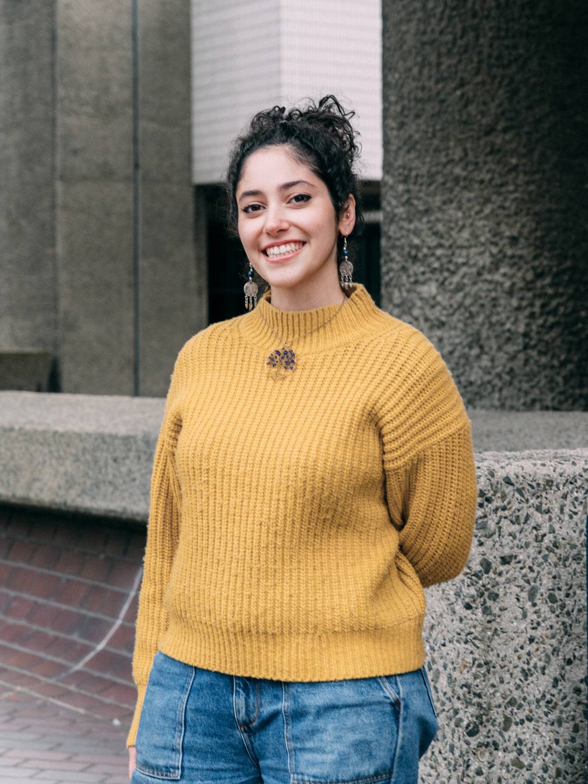 A photo of Riwa smiling and wearing a mustard-yellow sweater.
