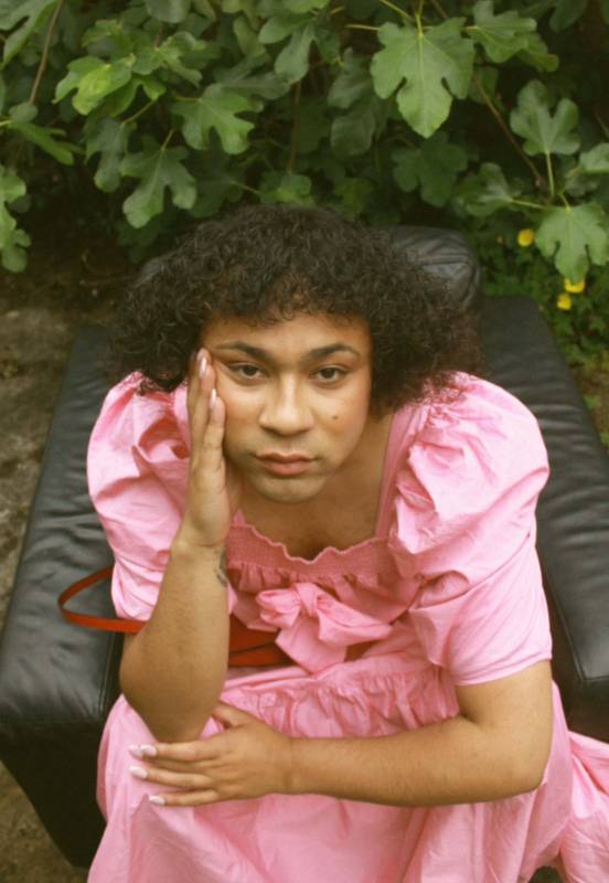 A photo of Travis in a pink dress. They are looking up, directly into the camera, with their right hand resting against their cheek. They are sitting in a black leather seat against some greenery.