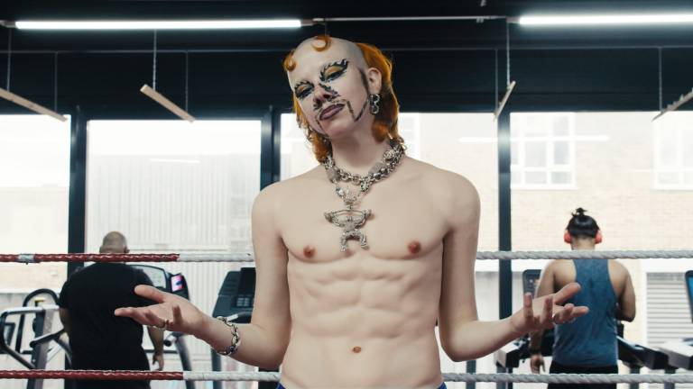 Wet Mess is white slim trans masc person. They stand in center frame, in a boxing ring in a gym, with two men on running machines behind them. They are topless, with big chains. They have two ginger horns and a receding hairline with a luscious ginger bob at the back. They have a handlebar moustache and detailed flame make up around their nose and eyes.