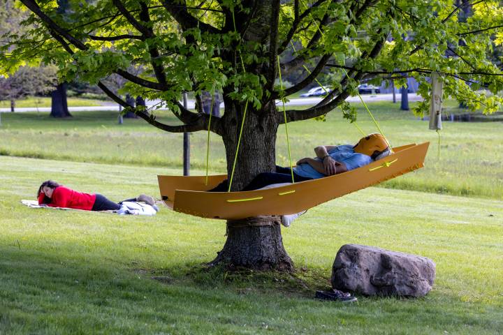 A person lies in a yellow hammock-style sculptural installation hanging from a leafy green tree. There is a mask over their face