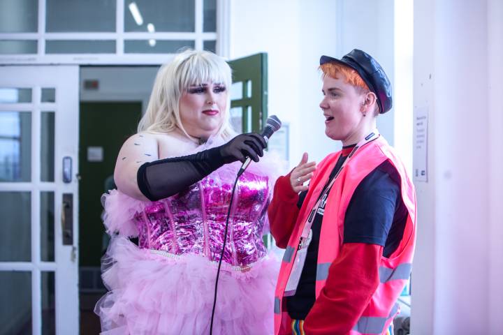 Drag performer Shepherd's Bush, dressed in a sparkly pink outfit, holds the microphone for an access support worker in a pink high-vis jacket to speak