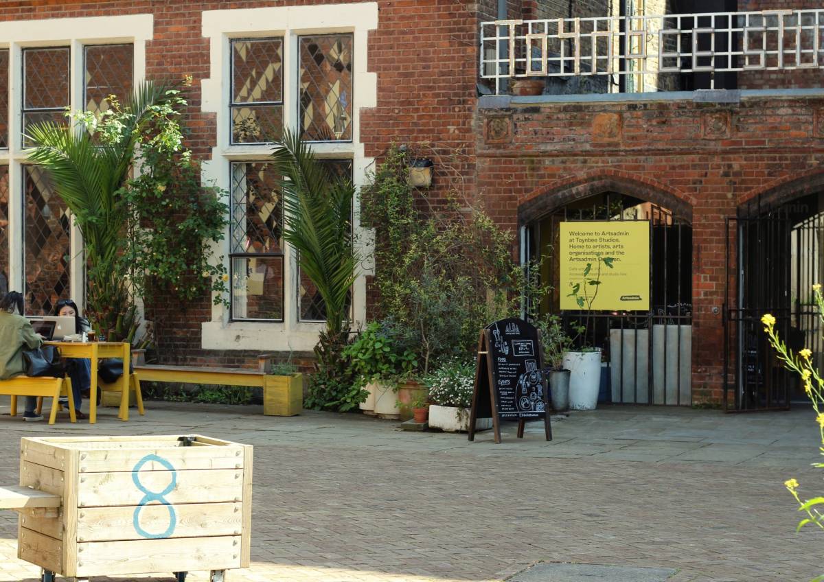 A photo of the outside of Toynbee Studios on a sunny day, with green plants, flower beds and some people working at a bench