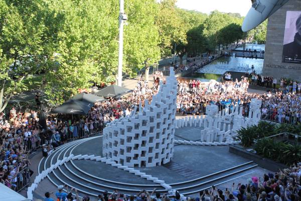 An image of the white domino sculptures piled up in a public space in Melbourne, with huge crowds surrounding them