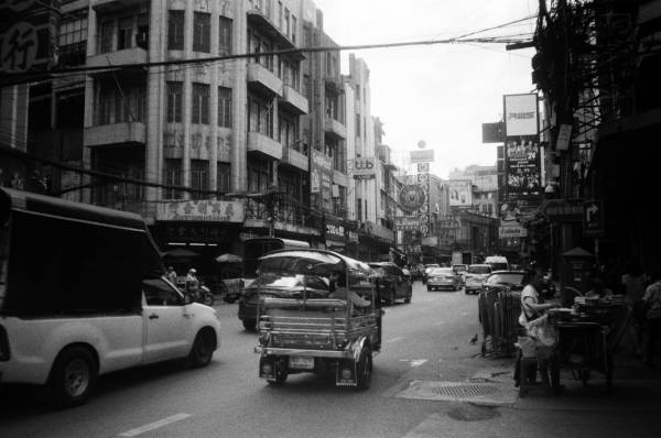 Black and white photo of a busy main road in China town, large advertisement signs on the sides of buildings, street food vendors along the pavements, tourists and locals mix.